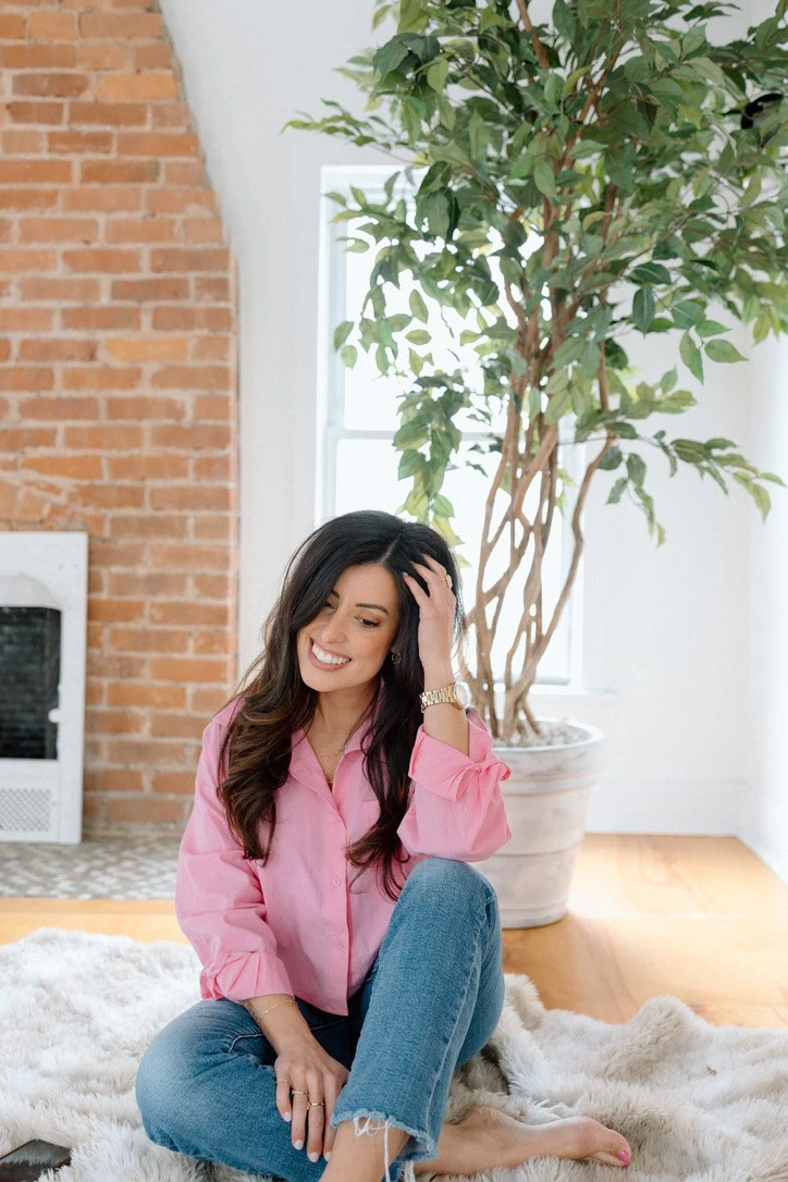Alana sitting on a rug at home in a pink shirt, smiling with a hand in her hair.
