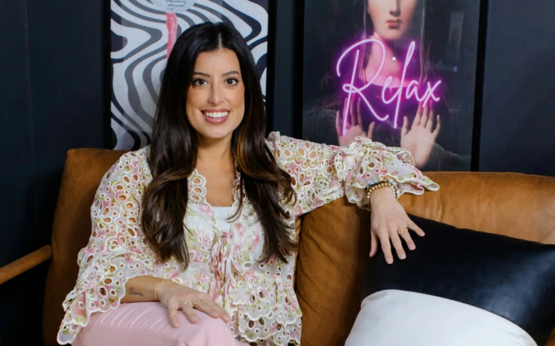 Alana on a leather sofa in her studio, a neon sign reading Relax behind her.