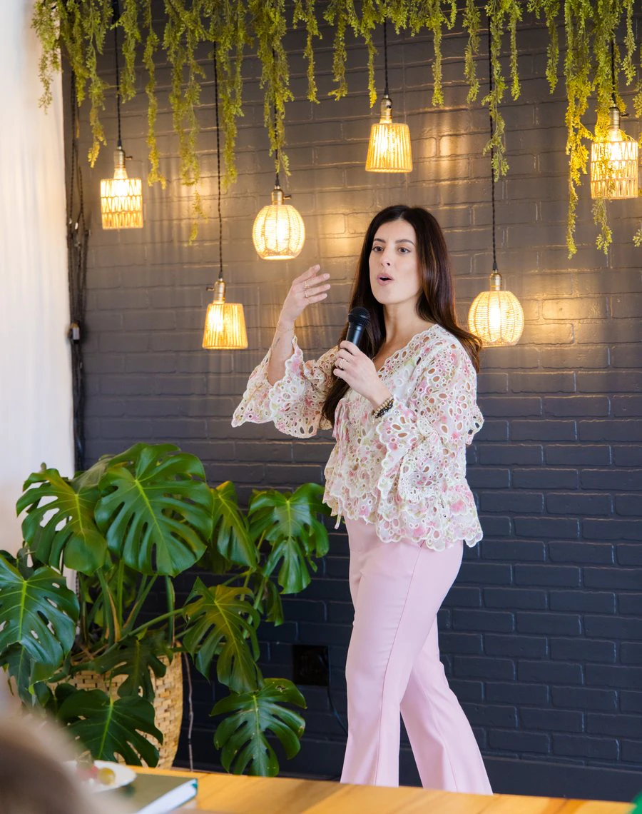 Alana speaking with a microphone beneath hanging greenery and warm pendant lamps.