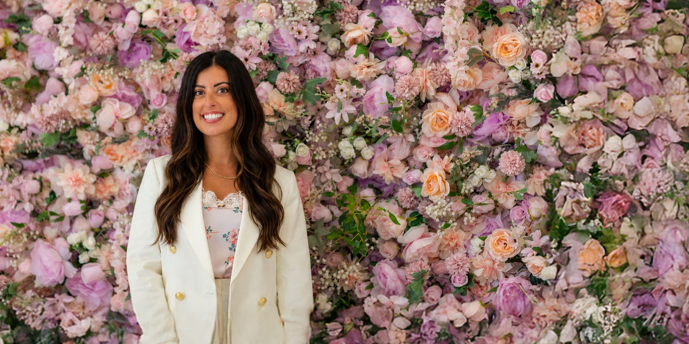 Alana Cummings in a white blazer in front of a floor-to-ceiling flower wall, smiling at the camera.